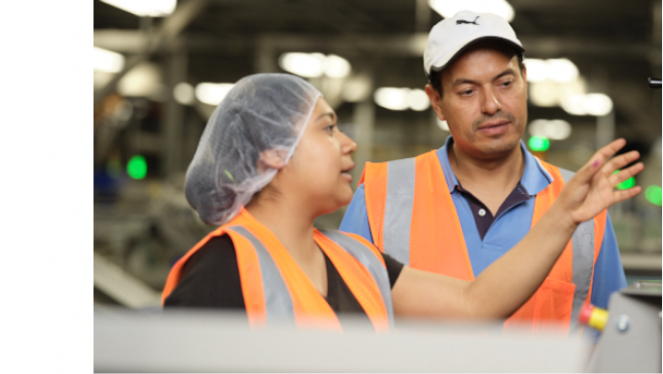 Two people wearing orange work vests are standing in a factory while the woman of the two wearing a hair net explains something
