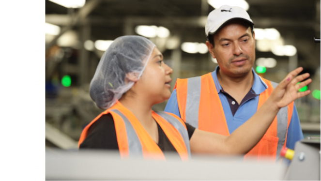 Two people wearing orange work vests are standing in a factory while the woman of the two wearing a hair net explains something