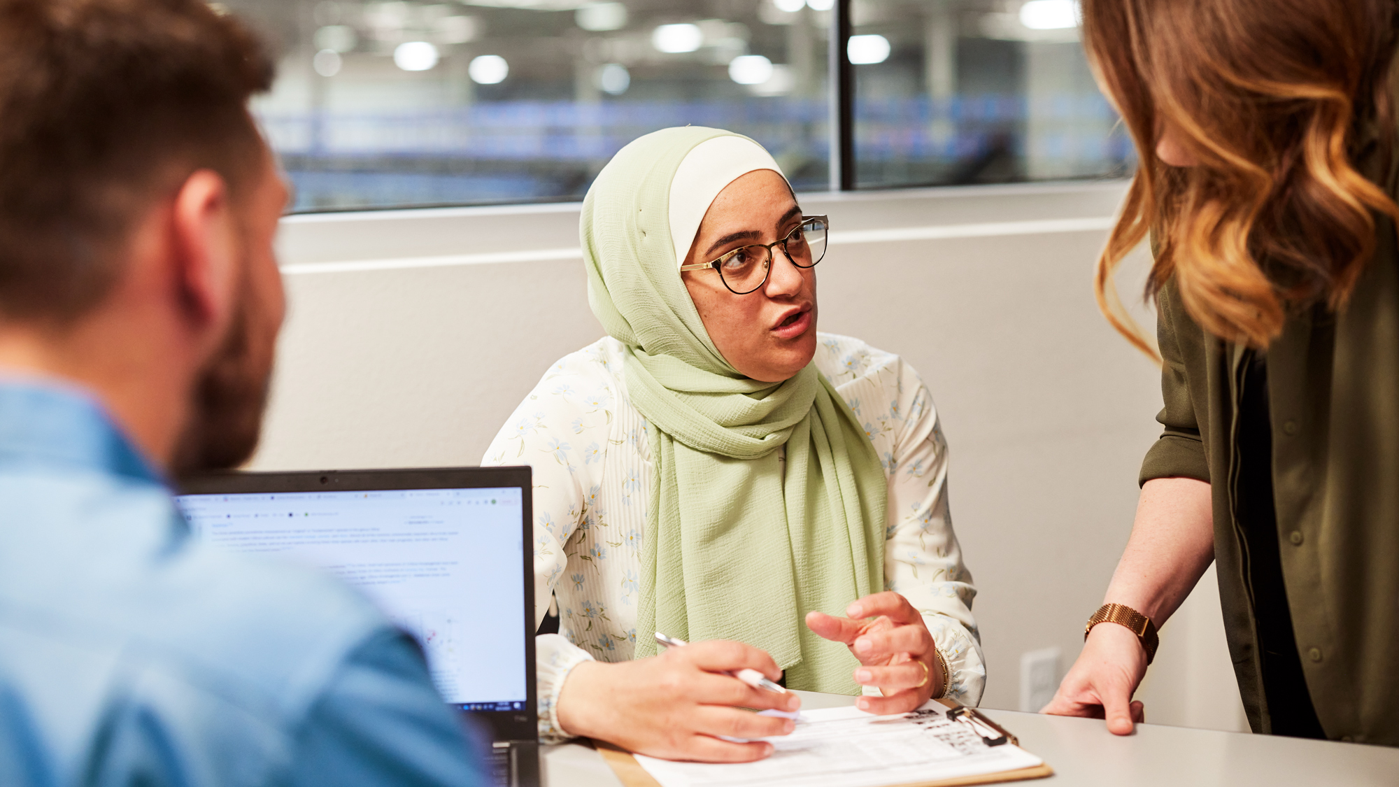 A woman wearing glasses and a hijab is sitting and explaining something to a sitting man and a standing woman at an office in front of a clipboard