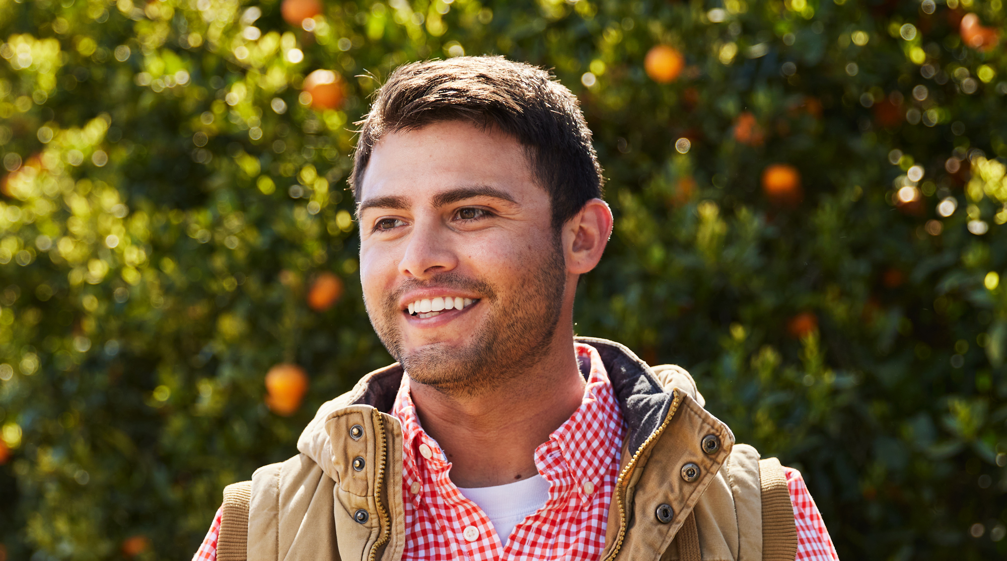 A man wearing a red flannel is standing in front of a tall bush of citrus and smiling