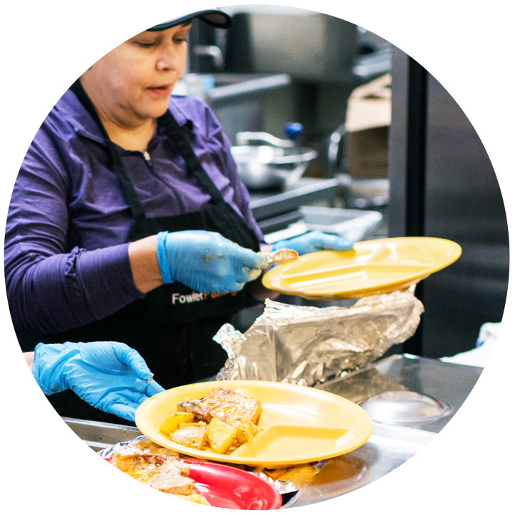 A woman wearing an apron and wearing gloves is spooning food into a plate in a cafeteria