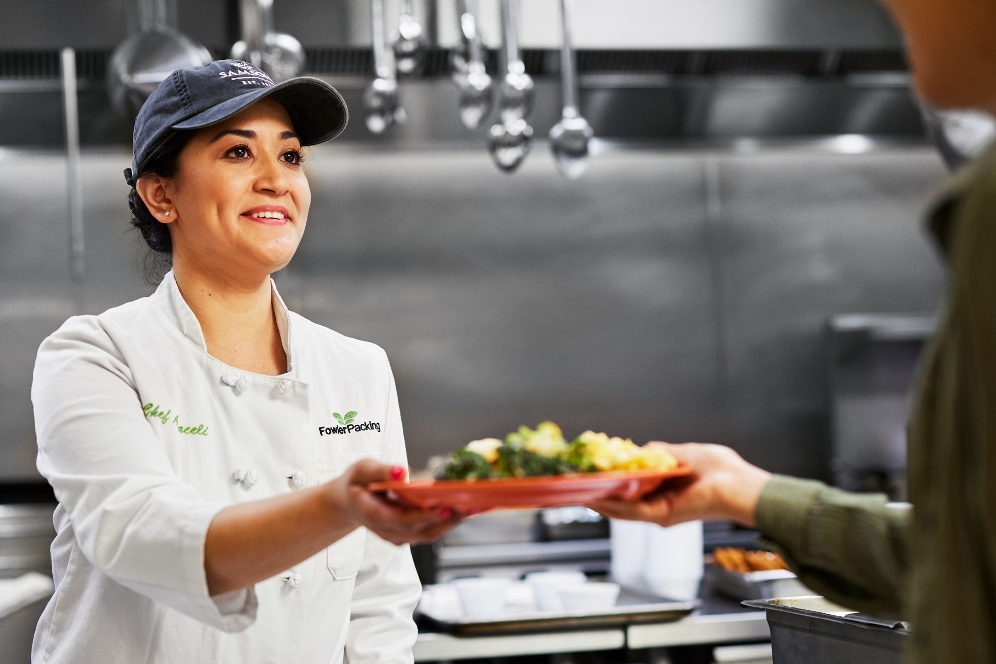 A female chef is wearing a chef apron and a navy cap is handing a plate of food to someone in a kitchen