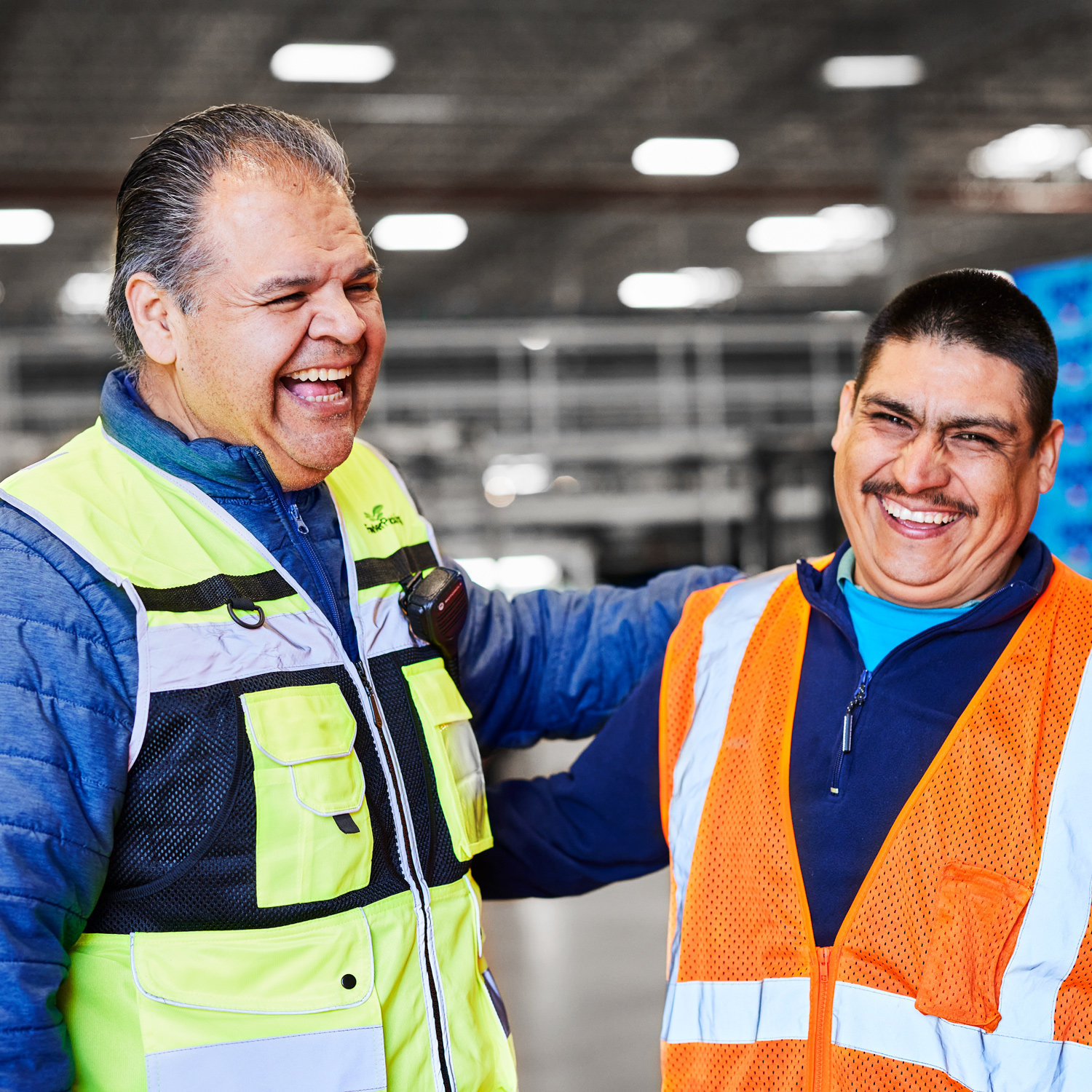 Two men wearing neon work vests are standing and laughing together in a factory