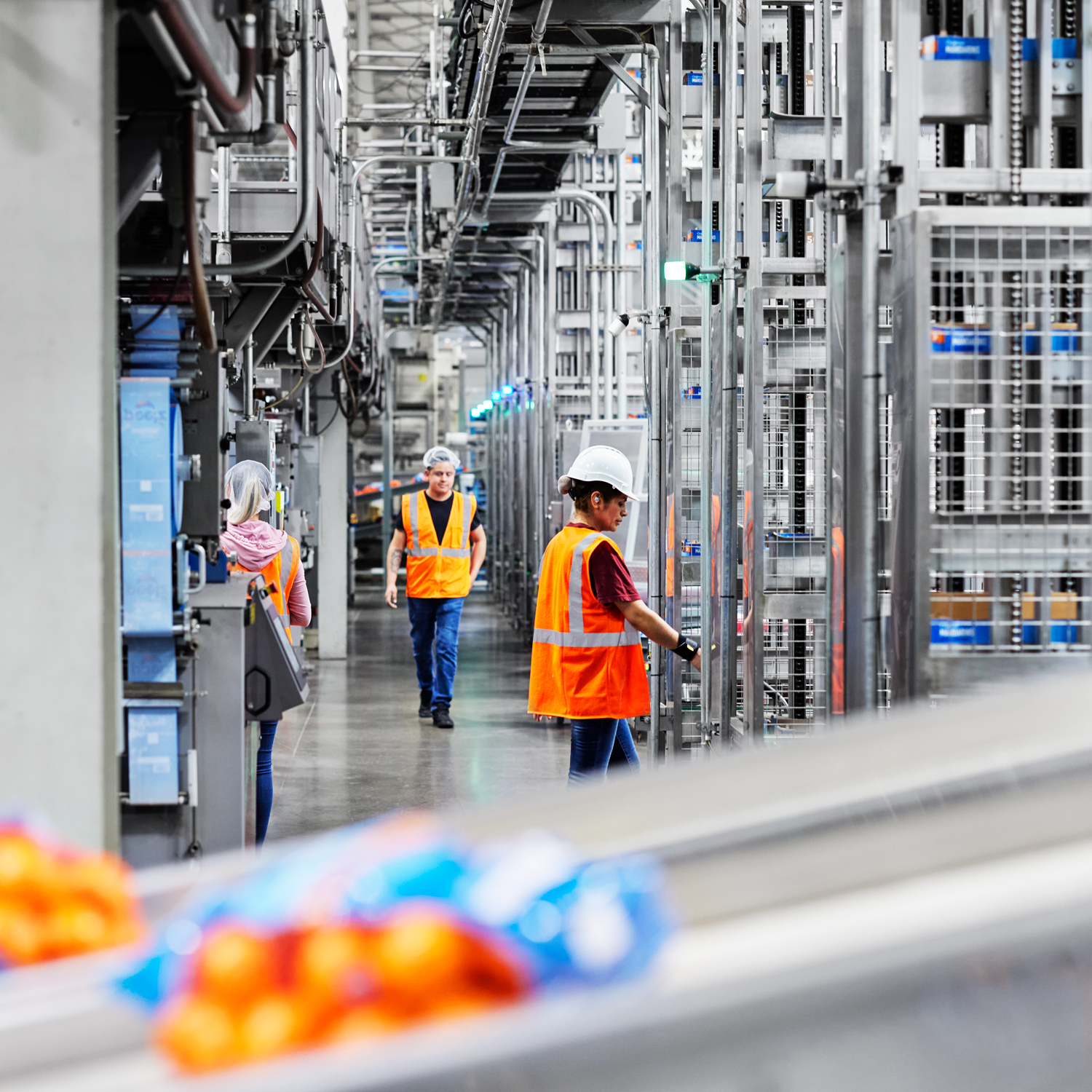 People in orange work vests are working on machines in a factory with mandarin orange peelz packages in the forefront